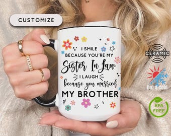 May include: White ceramic mug with a black handle and rim, featuring the text "I SMILE BECAUSE YOU'RE MY Sister In Law I LAUGH because you married MY BROTHER" with floral and star accents. The mug is labeled "CUSTOMIZE", "CERAMIC", "hot & cold", and "BPA FREE".