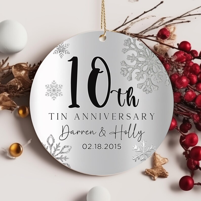 May include: A silver round Christmas ornament with a white background and silver glitter snowflakes. The ornament says "10th Tin Anniversary Darren & Holly 02.18.2015" in black text.