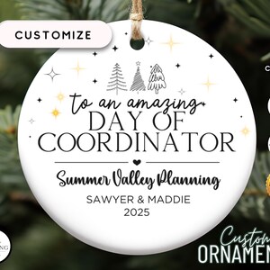 May include: White ceramic Christmas ornament with black text that reads "To an amazing Day of Coordinator Summer Valley Planning Sawyer & Maddie 2025". The ornament is decorated with gold stars and black and white illustrations of pine trees.