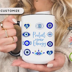 May include: White ceramic mug with a black handle and rim, featuring blue evil eye designs and the text "Protect your Energy" in blue script. The mug is held by a person with gold rings. The mug is labeled "Customize".