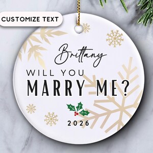 May include: A round white ceramic ornament featuring the text Brittany WILL YOU MARRY ME? and 2026 in black font. The design includes gold snowflake illustrations, a small green holly sprig with red berries, and a gold hanging cord.