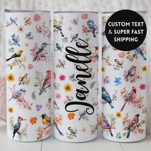 May include: A white tumbler featuring a colorful bird and floral design. The tumbler has a repeating pattern of various birds, pink and yellow flowers, and green leaves. The name "Janelle" is written in a cursive font on the tumbler.