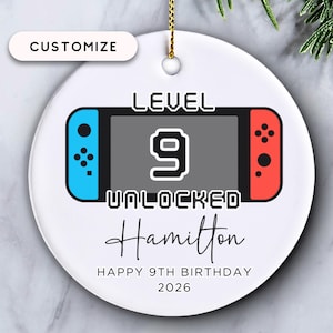 May include: White ceramic ornament with a video game controller design. The ornament features the text "LEVEL 9 UNLOCKED" and "Happy 9th Birthday 2026" with a name. The controller has blue and red accents.