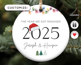 May include: A round white ceramic ornament featuring a string of colorful Christmas lights at the top. The text reads THE YEAR WE GOT ENGAGED 2025 with a red Santa hat on the zero. Below is the text Joseph & Harper and three small trees.