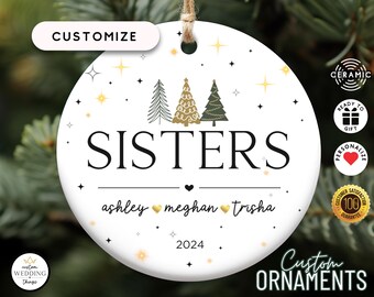May include: A round white ceramic ornament featuring the text SISTERS in black, three stylized Christmas trees, and the names ashley, meghan, and trisha separated by gold hearts. The year 2024 is printed at the bottom with gold and black stars.