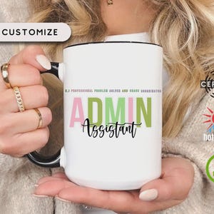 May include: White ceramic mug with black handle and rim, featuring the text "ADMIN Assistant" in pink and green. The mug also has the text "R.3 PROFESSIONAL PROBLEM SOLVER AND CHAIR COORDINATOR". The mug is held by a person.