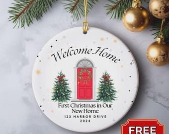 May include: A white ceramic Christmas ornament with the text "Welcome Home" and "First Christmas in Our New Home." It features a red door, Christmas trees, and the address "123 HARBOR DRIVE 2024."