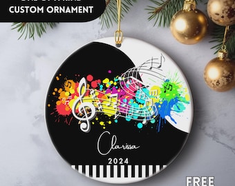 May include: A round, custom Christmas ornament with a musical theme. The ornament features a black and white design with colorful paint splatters, musical notes, and the name "Clarissa" with the year 2024. The text "ONE OF A KIND CUSTOM ORNAMENT" is at the top.