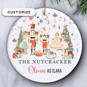 May include: A white ceramic Christmas ornament with a gold string. The ornament features a colorful illustration of characters from "The Nutcracker" ballet, including a nutcracker, mouse, and Clara. Text includes "The Nutcracker" and "Olivia as Clara".