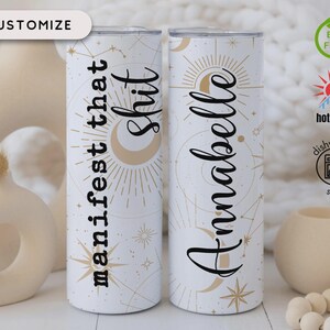 May include: Two white stainless steel tumblers with celestial designs. One tumbler reads "manifest that shit" and the other reads "Anabelle" in black script. The tumblers are decorated with gold stars, moons, and sunbursts. The tumblers are BPA free, dishwasher safe, and suitable for hot or cold beverages.