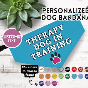 May include: A personalized dog bandana in turquoise blue with the text "THERAPY DOG IN TRAINING" in white. A pink paw print is at the bottom. The bandana has a black adjustable strap and the option to customize the text.