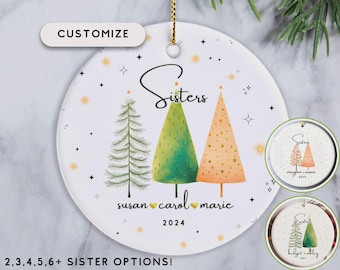 May include: A white ceramic Christmas ornament with a gold string. It features three stylized Christmas trees in green and orange, the word "Sisters", names, and the year 2024. The text "Customize" is also visible.