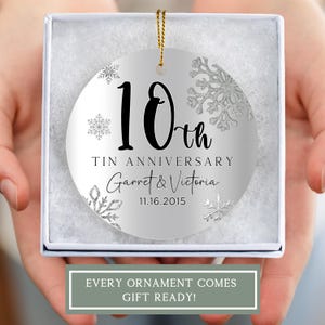 May include: A silver round Christmas ornament with a white background and silver glitter snowflakes. The ornament features black text that reads "10th Tin Anniversary Garret & Victoria 11.16.2015".