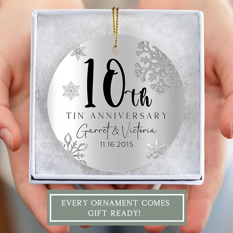 May include: A silver round Christmas ornament with a white background and silver glitter snowflakes. The ornament features black text that reads "10th Tin Anniversary Garret & Victoria 11.16.2015".
