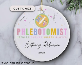 May include: A round white ceramic ornament featuring the text PHLEBOTOMIST in colorful letters, with the subtext NEEDLES. SCRUBS. RUBBER GLOVES. It includes a circular graphic with a syringe, the letters CPT, the name Bethany Robinson, and 2026.