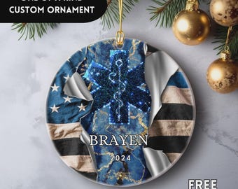 Può includere: Ornamento rotondo personalizzato con un design marmorizzato blu e oro, con un simbolo blu a stella della vita. L'ornamento ha il nome "Brayen" e l'anno "2024" stampati sopra. Lo sfondo mostra una bandiera americana.