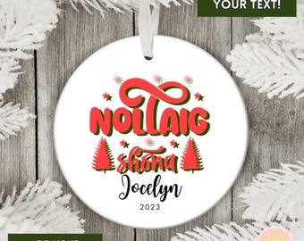 May include: White ceramic Christmas ornament with red and green text that reads "Nollaig Shona Jocelyn 2023". The ornament has a white ribbon for hanging and features Christmas tree and snowflake graphics.