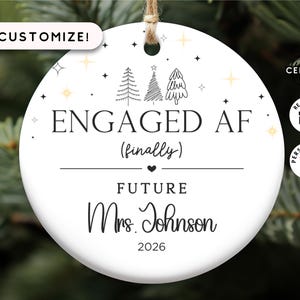 May include: White ceramic Christmas ornament with a black and white illustration of three Christmas trees and the text "ENGAGED AF (finally) FUTURE Mrs. Johnson 2026".