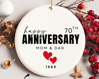 Può includere: Ornamento a disco in ceramica bianca con cordino dorato, con la scritta "happy ANNIVERSARY" in nero, con "70th" sopra. Sotto, si legge "MOM & DAD" e "1989" con due cuori rossi. Le parole "ADD PERSONALIZATIONS" sono in basso.