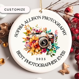 May include: A white ceramic ornament with a watercolor design of a camera surrounded by colorful flowers. The ornament has the text "SOPHIA ALLISON PHOTOGRAPHY BEST PHOTOGRAPHER EVER 2025". A gold string is attached for hanging.