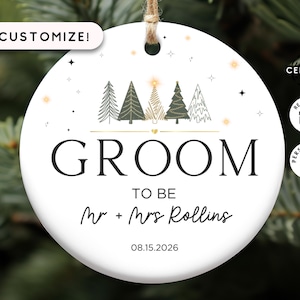 May include: A round white ceramic ornament featuring a line of five stylized Christmas trees above the text GROOM TO BE, Mr + Mrs Rollins, and the date 08.15.2026. The design includes small gold stars and a jute hanging string.