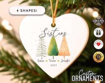 May include: A heart-shaped ceramic ornament with the word "Sisters" in script. It features three illustrated Christmas trees in green and orange, with names and the year 2027. The ornament has a gold string for hanging.