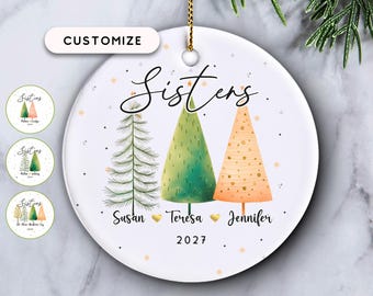 May include: A round white ceramic ornament featuring the word Sisters in black script. Below are three stylized Christmas trees in green and peach tones, labeled with the names Susan, Teresa, and Jennifer, separated by gold hearts and the year 2027.