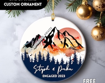 Può includere: Ornamento rotondo in ceramica bianca con un design di montagna e tramonto. Il testo "OUR ADVENTURE AWAITS" è sopra le montagne, con "Steph & Parker ENGAGED 2023" sotto. L'ornamento è personalizzato e unico.