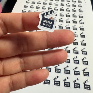 May include: A sheet of white paper with multiple black and white clapperboard stickers. The stickers are arranged in rows and columns. A hand is holding one of the stickers up to the camera.