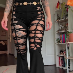 May include: Black high-waisted flared pants featuring intricate cut-out lattice detailing along the thighs and legs, paired with a dark green sleeveless top with a front lace-up design and a gold-toned coin chain belt.