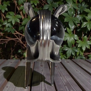 Puede incluir: Una escultura brillante de color cromo de un animal estilizado, posiblemente un conejo o una liebre, con largas orejas y un cuerpo redondeado. La escultura tiene una superficie reflectante y se apoya sobre tres patas. El fondo presenta follaje verde y una terraza de madera.