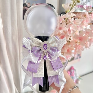 May include: A decorative light stick with a clear orb and black handle. The light stick is adorned with white and lavender ribbons, faux pearl accents, and small white wings. A large purple jewel is centered on the bow.