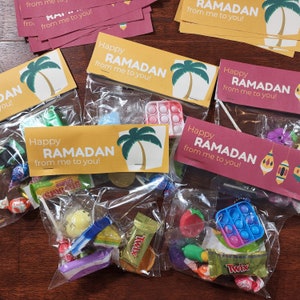 May include: Four clear plastic bags filled with small toys and candy. Each bag has a colorful tag with the text "Happy Ramadan from me to you!" and a palm tree graphic.