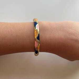 Könnte beinhalten: Ein goldenes Armreif-Armband mit einem geometrischen Muster aus rosa, orange und blauem Emaille.