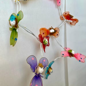 Pode incluir: Uma sequência de luzes decorativas com figuras de fadas caprichosas. As fadas têm asas coloridas, incluindo rosa, amarelo, verde, laranja e roxo, e rostos de boneca. As luzes são enfiadas em um fio branco.