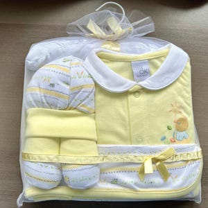 Könnte beinhalten: Ein gelbes Baby-Outfit-Set mit weißem Kragen, Mütze und Lätzchen. Das Outfit hat ein kleines gesticktes Design eines Küken auf der Vorderseite. Das Set ist in einer durchsichtigen Plastiktüte mit einem weißen Band verpackt.