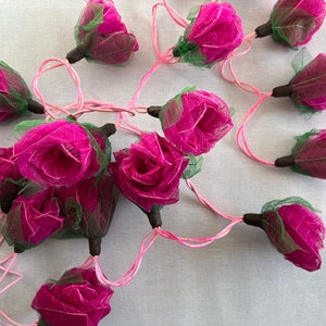 May include: A string of pink and green paper rose lights. The roses are made of delicate, translucent paper and are strung together with pink wire.
