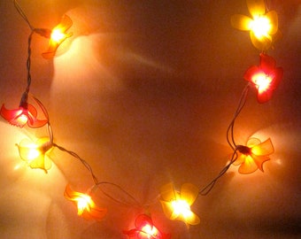 Puede incluir: Cadena de luces con bombillas en forma de flor en tonos amarillos, naranjas y rojos. Las luces están enhebradas en un círculo.