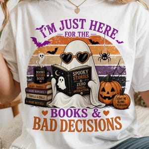 Könnte beinhalten: Ein weißes T-Shirt mit einer Grafik eines Geistes, der herzförmige Sonnenbrillen trägt und ein Buch mit dem Titel Spooky Stories & Zero Social Interaction liest. Das Design enthält Kürbisse, Fledermäuse, eine Spinne und den Text I'm Just Here For The Books & Bad Decisions.