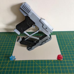 Pode incluir: Uma pistola de brinquedo cinza e preta &eacute; exibida em uma superf&iacute;cie reflexiva, com seu reflexo abaixo. A pistola apresenta um design detalhado, incluindo um topo canelado e uma empunhadura texturizada. Dois pequenos objetos espinhosos coloridos.
