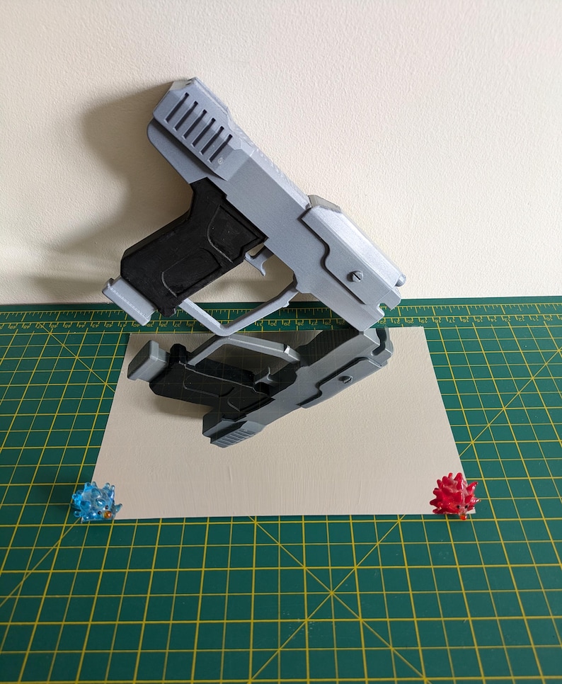 Pode incluir: Uma pistola de brinquedo cinza e preta &eacute; exibida em uma superf&iacute;cie reflexiva, com seu reflexo abaixo. A pistola apresenta um design detalhado, incluindo um topo canelado e uma empunhadura texturizada. Dois pequenos objetos espinhosos coloridos.