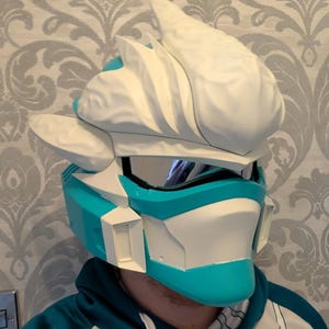 Pode incluir: Um capacete futurista branco e azul-turquesa, com viseira e design angular. O capacete tem uma estrutura &uacute;nica, em camadas, com uma crista proeminente no topo. O esquema de cores &eacute; um contraste marcante de azul-turquesa brilhante e branco.