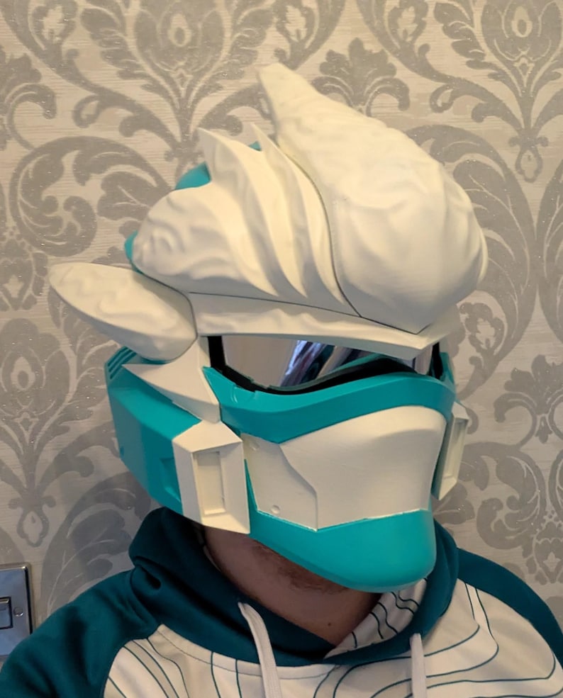 Pode incluir: Um capacete futurista branco e azul-turquesa, com viseira e design angular. O capacete tem uma estrutura &uacute;nica, em camadas, com uma crista proeminente no topo. O esquema de cores &eacute; um contraste marcante de azul-turquesa brilhante e branco.
