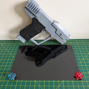 Pode incluir: Uma pistola de brinquedo impressa em 3D em cinza claro e preto, com um design detalhado. A pistola de brinquedo est&aacute; colocada sobre uma superf&iacute;cie preta reflexiva, com dois pequenos objetos decorativos coloridos em cada lado.