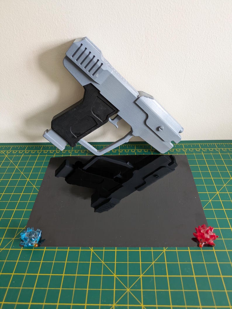 Pode incluir: Uma pistola de brinquedo impressa em 3D em cinza claro e preto, com um design detalhado. A pistola de brinquedo est&aacute; colocada sobre uma superf&iacute;cie preta reflexiva, com dois pequenos objetos decorativos coloridos em cada lado.