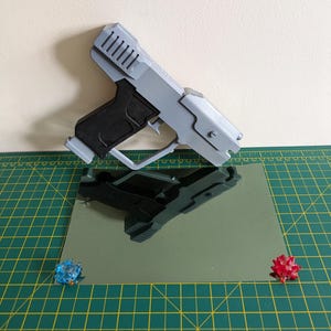 Pode incluir: Uma pistola de brinquedo cinza e preta com um design futurista. A arma de brinquedo est&aacute; sobre uma superf&iacute;cie refletora, com dois pequenos objetos decorativos coloridos em cada lado. O fundo &eacute; bege claro.