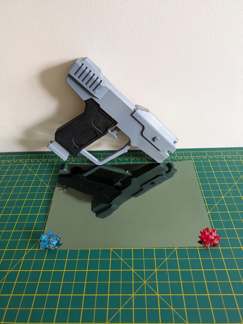 Pode incluir: Uma pistola de brinquedo cinza e preta com um design futurista. A arma de brinquedo est&aacute; sobre uma superf&iacute;cie refletora, com dois pequenos objetos decorativos coloridos em cada lado. O fundo &eacute; bege claro.