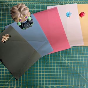 May include: An assortment of colorful envelopes in shades of green, blue, pink, white, and gold. Each envelope is adorned with a decorative embellishment, including a gold bee and small floral accents. A figurine is placed on top of the envelopes.