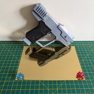 Pode incluir: Uma pistola de brinquedo cinza e preta com um design detalhado, repousando sobre uma superf&iacute;cie bege. A pistola &eacute; refletida na superf&iacute;cie. Dois pequenos objetos decorativos coloridos s&atilde;o colocados nos cantos da superf&iacute;cie bege.
