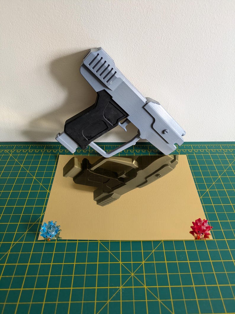 Pode incluir: Uma pistola de brinquedo cinza e preta com um design detalhado, repousando sobre uma superf&iacute;cie bege. A pistola &eacute; refletida na superf&iacute;cie. Dois pequenos objetos decorativos coloridos s&atilde;o colocados nos cantos da superf&iacute;cie bege.